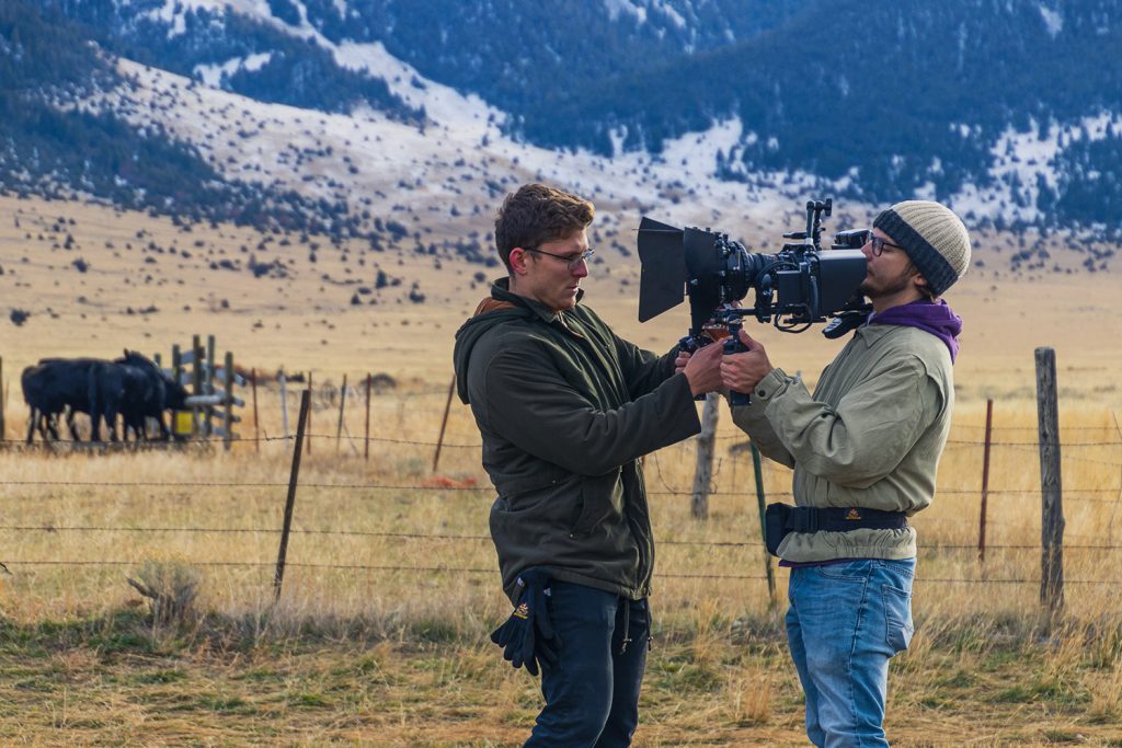 Two camera operators filming in a field below mountains